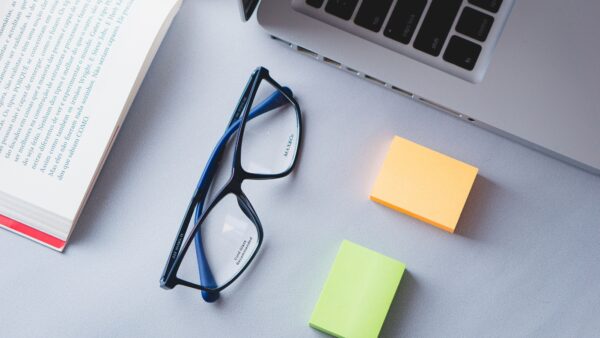 Book, laptop, eyeglasses, and sticky notes on a desk, representing learning and skill development.