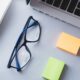 Book, laptop, eyeglasses, and sticky notes on a desk, representing learning and skill development.